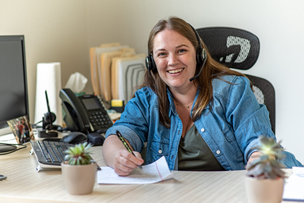 Kayte at her desk, smiling at the camera, writing on a piece of paper