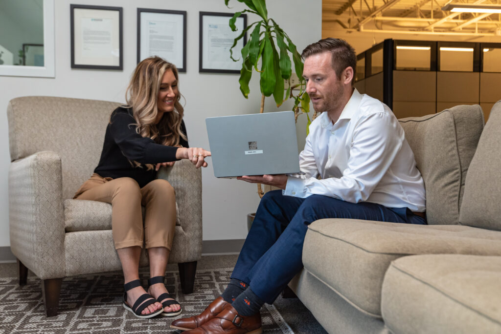 Man on a couch holding a laptop with a woman on a chair next to him pointing to his laptop screen